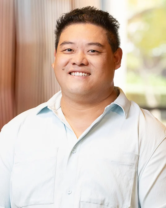 Dr Jay Gong, smiling, wearing a light blue shirt, in a warm clay-toned interior